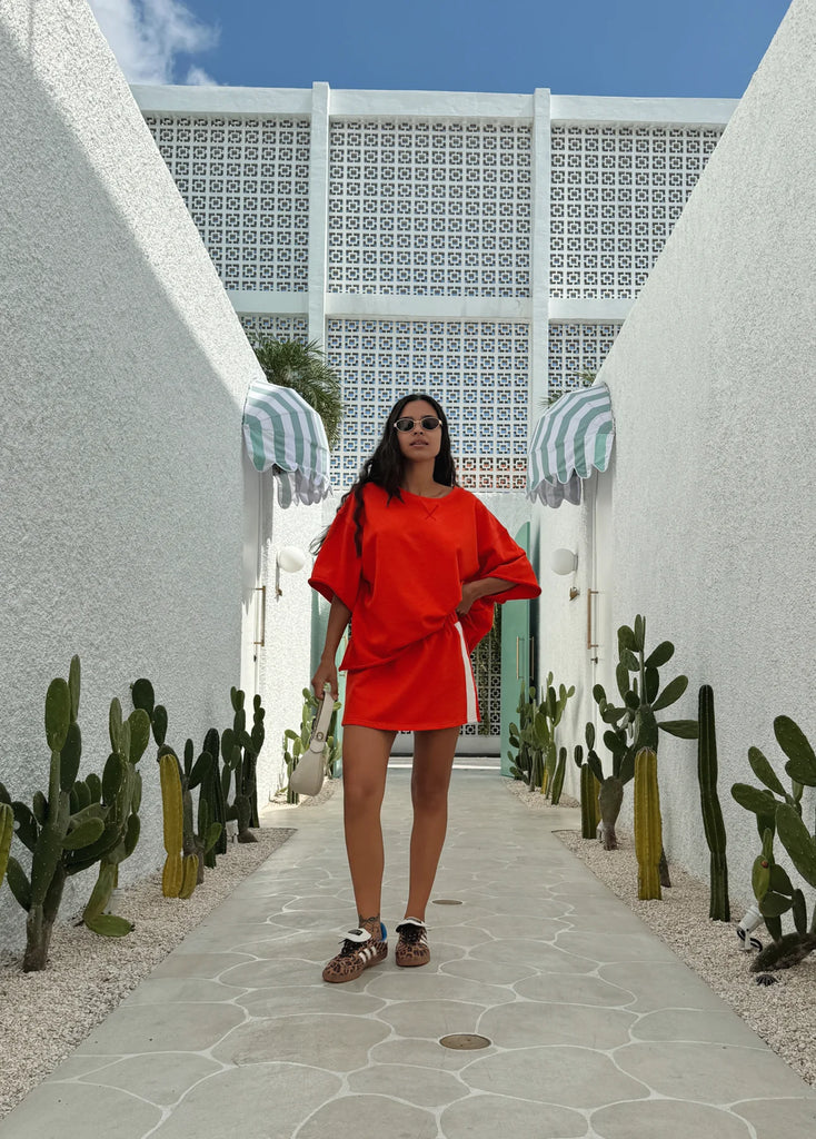 Woman in a red outfit standing in a sunlit outdoor setting with cacti and a patterned wall.